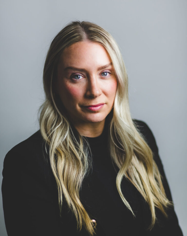 A woman with long blonde hair wearing a black jacket poses against a plain, light gray background.