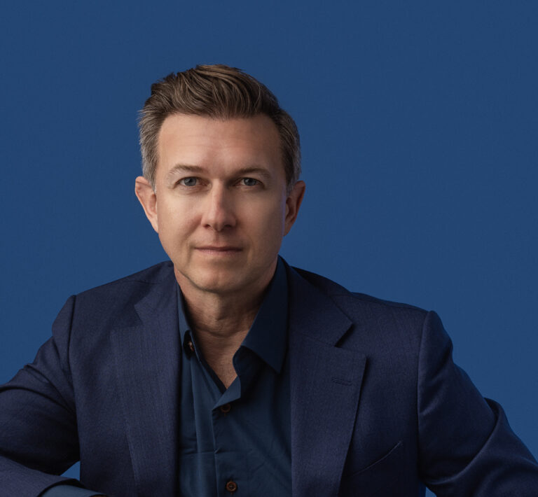 A man in a navy suit and matching shirt poses confidently against a blue background. His expression is calm and composed, conveying professionalism.